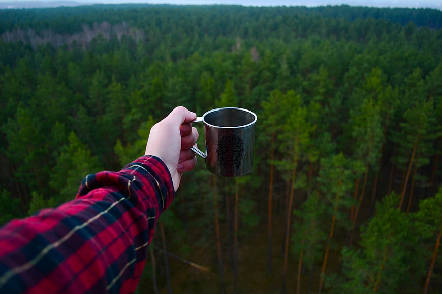 Hand holding metal camping mug over pine forest view