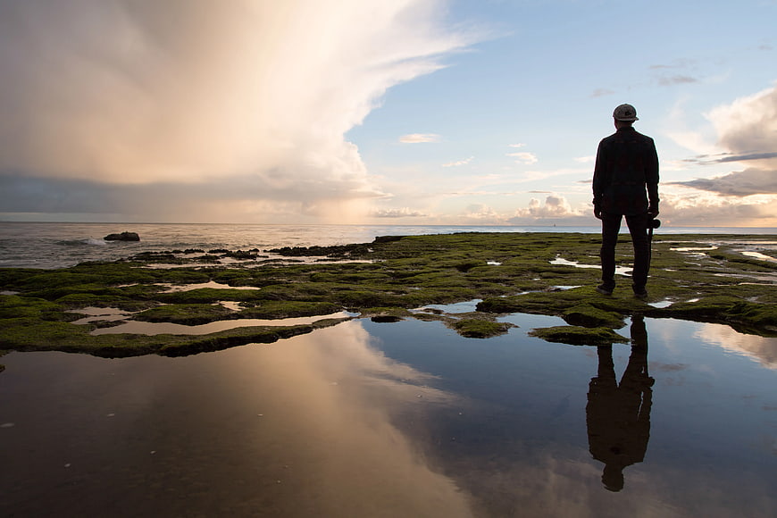 Silhouette of Man Standing on Mossy Rocks by Ocean at Sunset