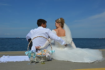 Wedding couple seated on pier with picnic basket and champagne by the sea