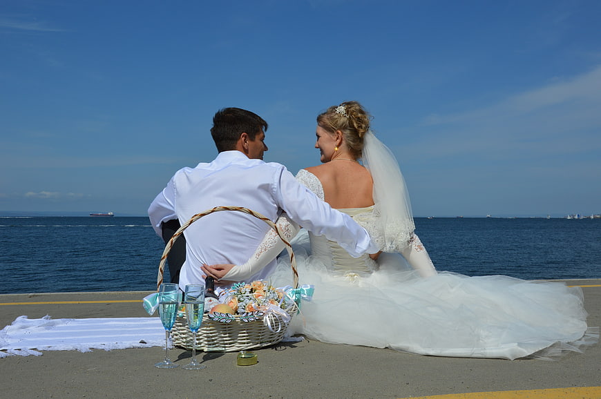 Wedding couple seated on pier with picnic basket and champagne by the sea