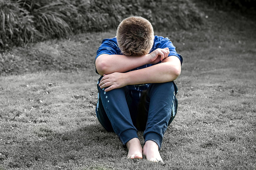Sad young boy sitting alone on grass with head down