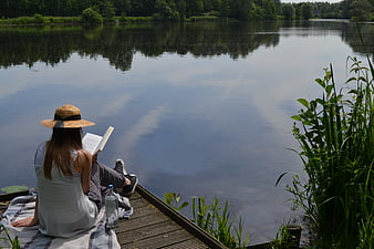 Woman reading book on wooden dock by calm lake in summer