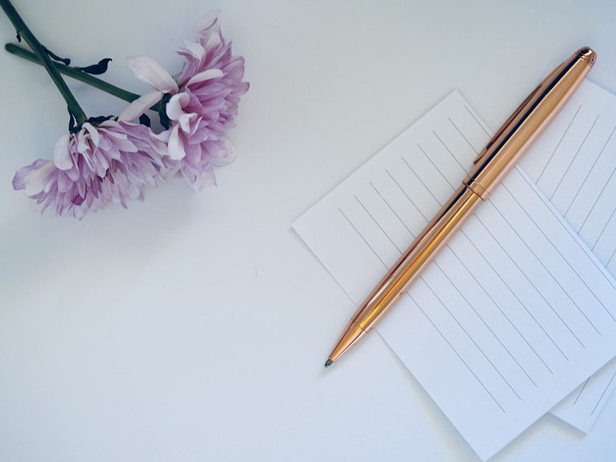Minimalist flat lay with gold pen, lined paper, and pink flowers on white background