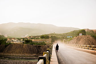 Lone figure walking on a dusty bridge in a rural mountain landscape