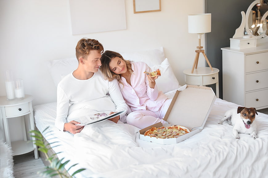 Happy couple eating pizza in bed with dog