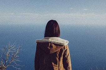 Woman in brown jacket gazing at calm blue ocean from cliff
