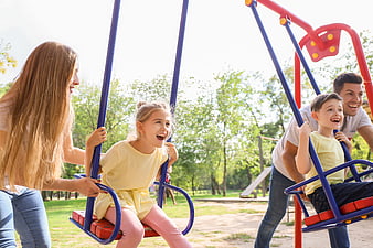 Happy family pushing children on swings at sunny park