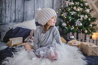 Little girl in knit hat sitting by Christmas tree with gifts
