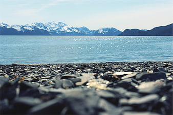 Low angle view of rocky beach with sparkling blue water and snow-capped mountains