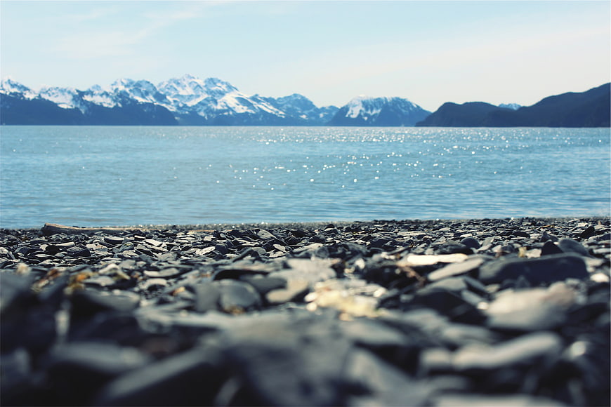 Low angle view of rocky beach with sparkling blue water and snow-capped mountains