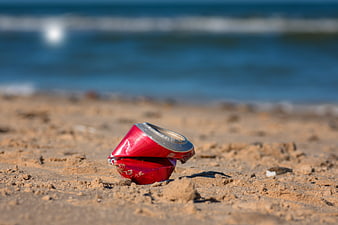 Crushed red aluminum can on sandy beach with ocean background