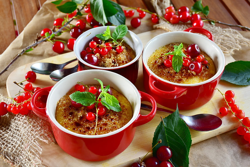 Three red ramekins of creme brulee with fresh cherries and mint