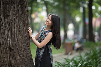 Smiling Asian Woman Leaning Against Large Tree Trunk in Park