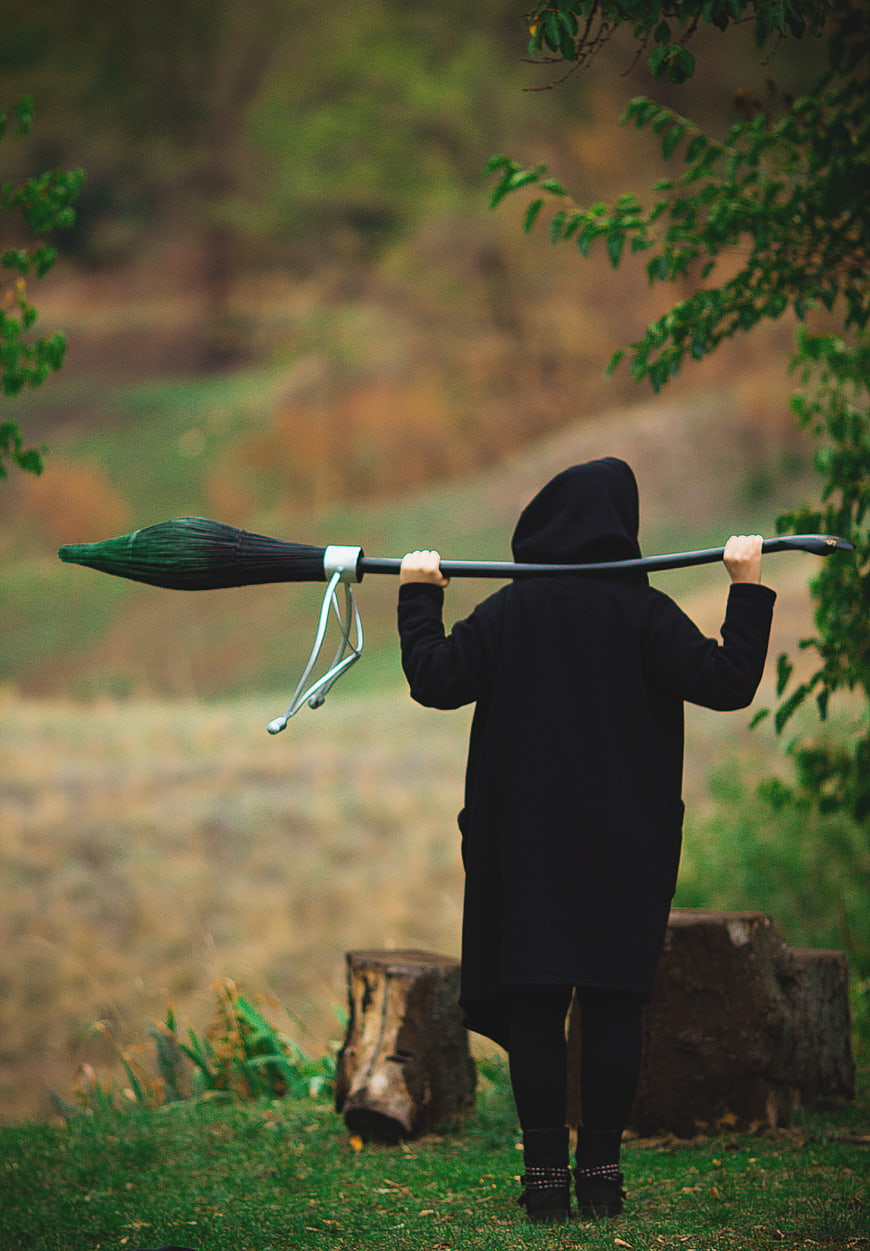 Person in black hooded robe carrying a broomstick in a forest