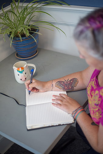 Woman writing in notebook with unicorn mug and tattoo