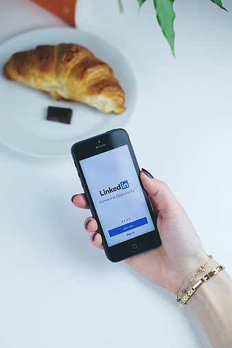Hand holding smartphone with LinkedIn app and croissant