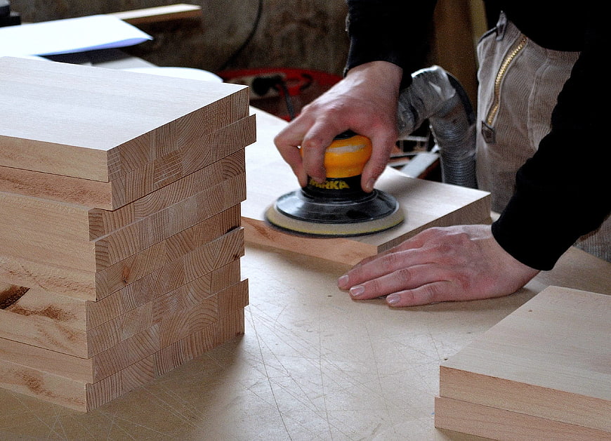 Carpenter Using Orbital Sander On Wood Planks In Workshop