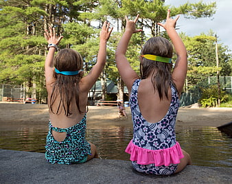 Two Girls in Swimsuits Sitting by Lake with Hands Raised