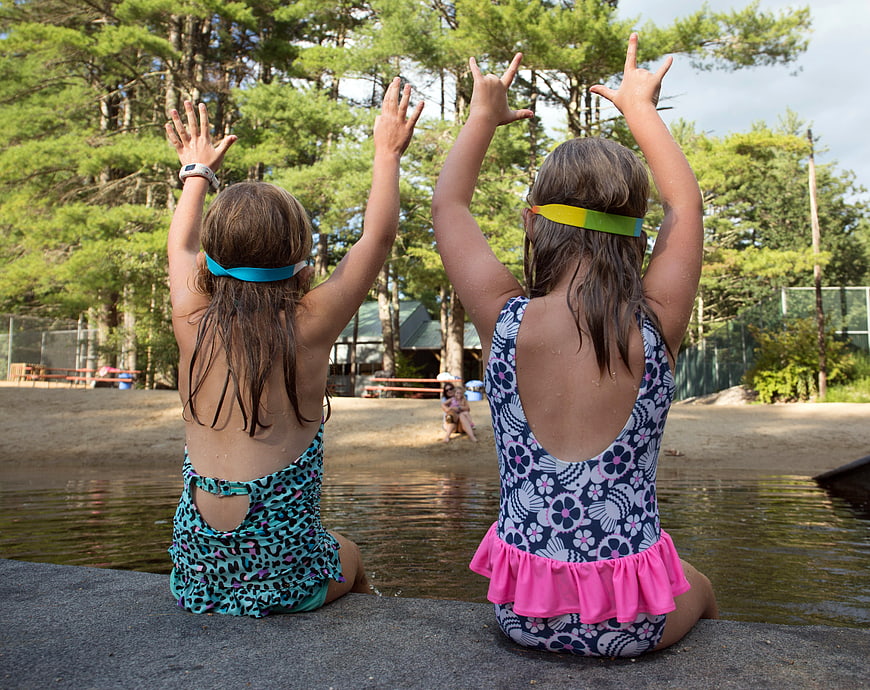 Two Girls in Swimsuits Sitting by Lake with Hands Raised