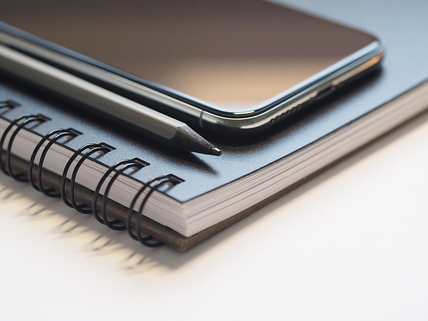 Close-up of smartphone, pencil, and spiral notebook on white desk