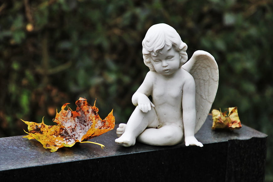 White Angel Statue Sitting on Stone with Autumn Leaves