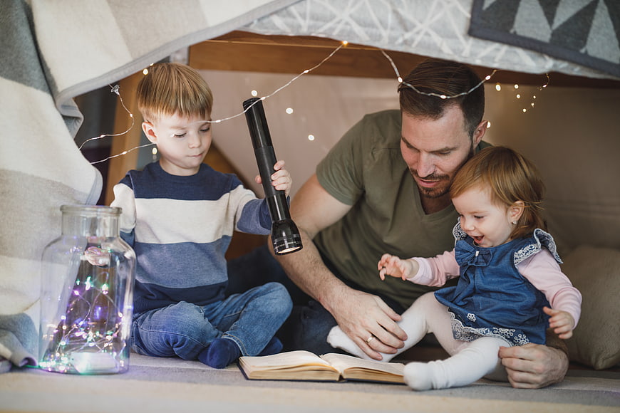Father reading to children in cozy blanket fort with fairy lights