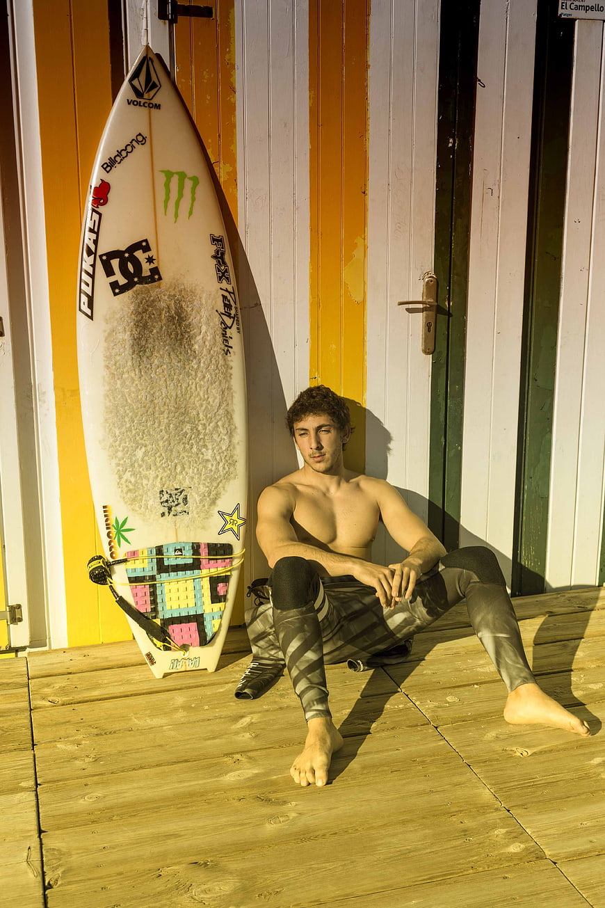 Young Surfer Sitting on Wooden Deck with Surfboard