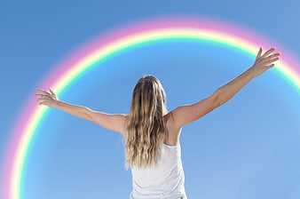 Woman with arms raised embracing a rainbow against blue sky