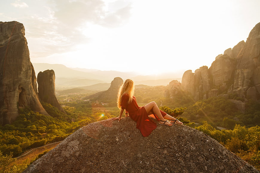 Woman in Red Dress Sitting on Rock Overlooking Meteora at Sunset
