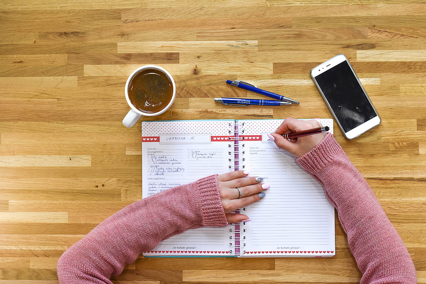 Woman writing in notebook on wooden desk with coffee and smartphone