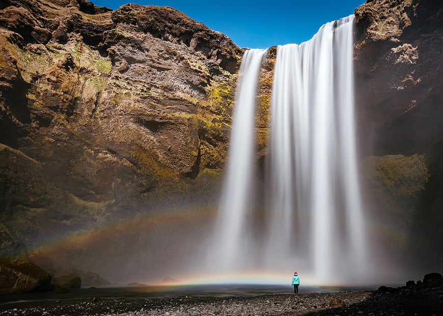 Lone Hiker Standing Before Majestic Waterfall with Rainbow