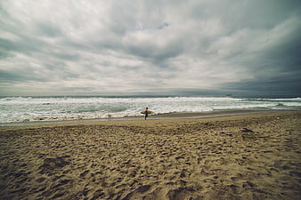 Lone Surfer Walking on Sandy Beach Under Cloudy Sky