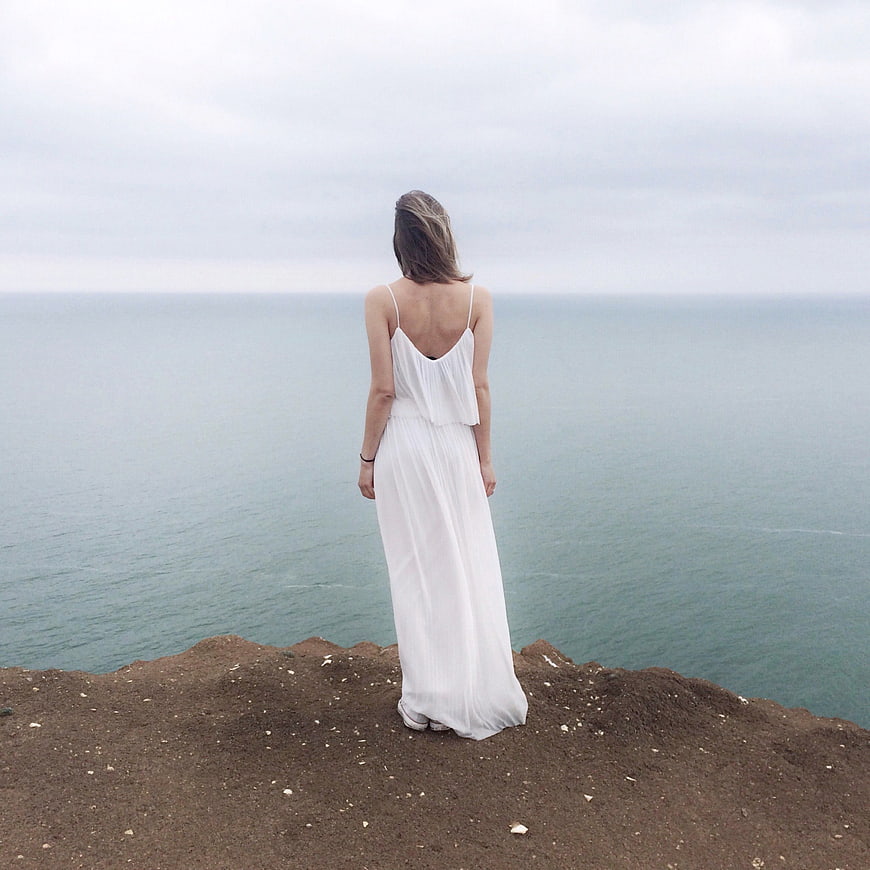 Woman in White Dress Standing on Cliff Overlooking Ocean