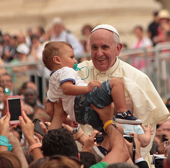 Pope Francis smiling while holding a baby in crowd