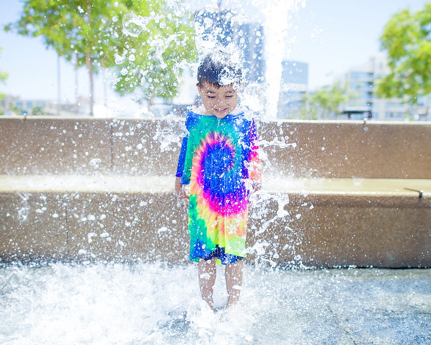 Happy Boy Playing in Water Fountain Wearing Tie-Dye Shirt