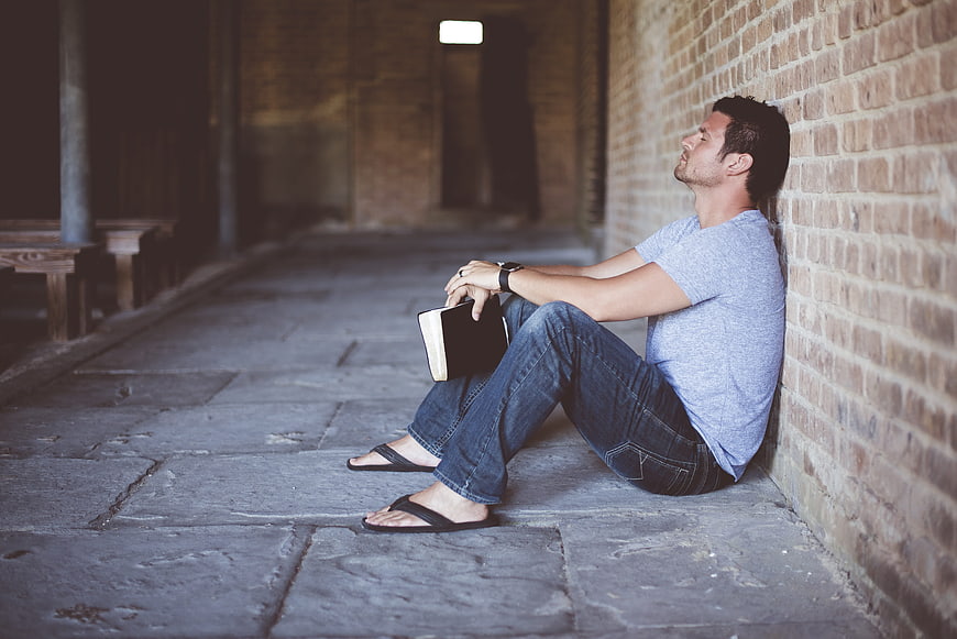 Man sitting against brick wall holding book in contemplation