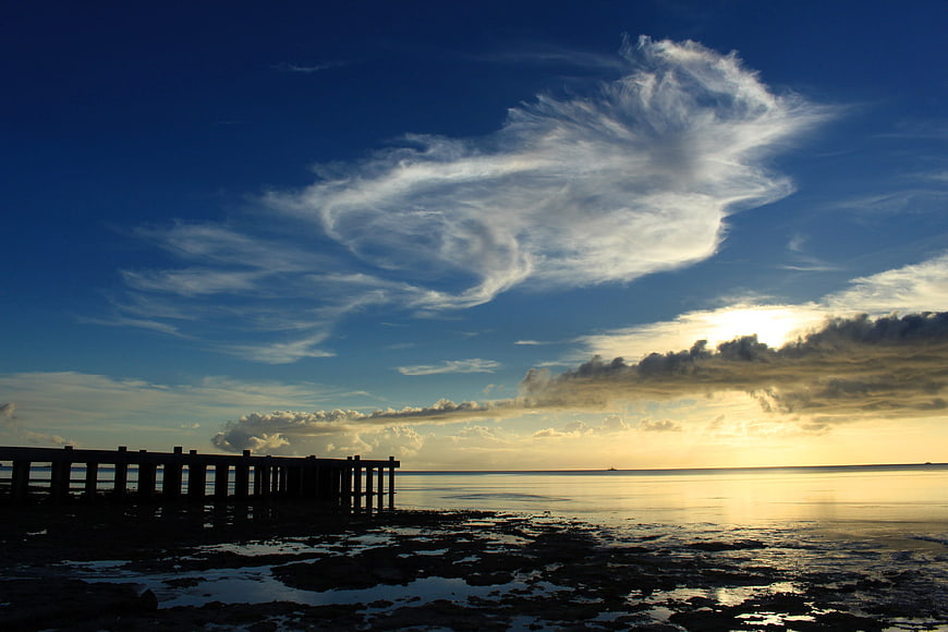 Silhouetted pier at sunset with dramatic clouds over calm ocean