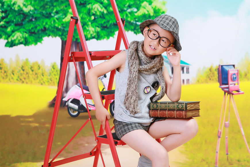 Young boy with glasses and hat sitting on red ladder with books