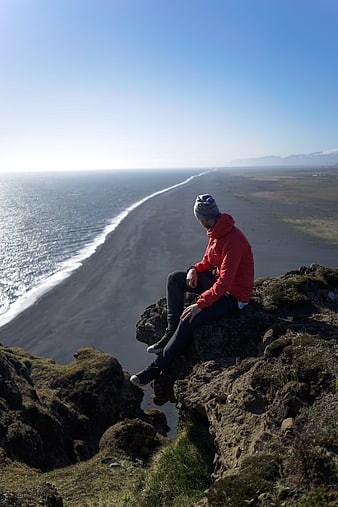 Man in red jacket sitting on cliff overlooking black sand beach