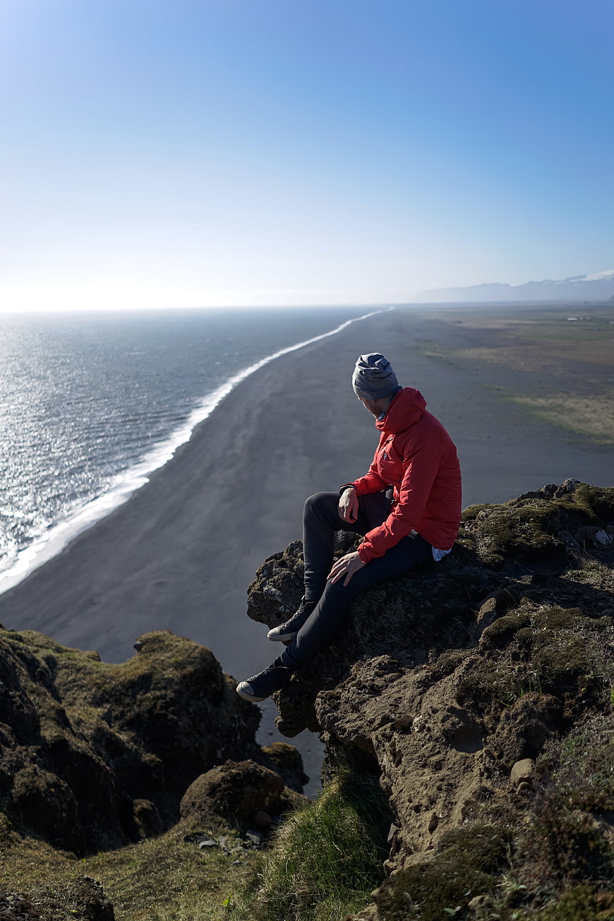 Man in red jacket sitting on cliff overlooking black sand beach