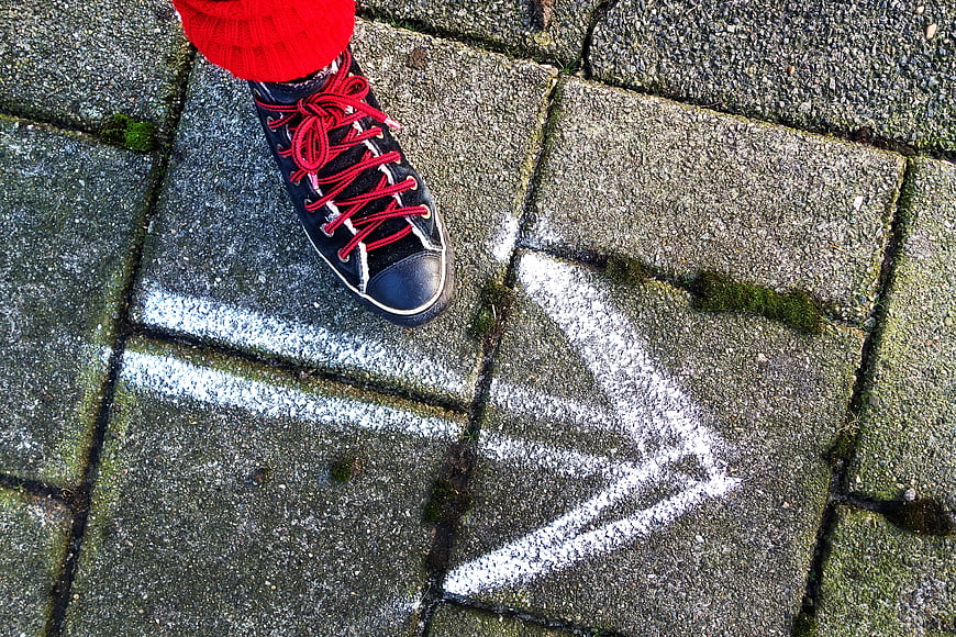 Black sneaker with red laces stepping on pavement with white chalk arrow