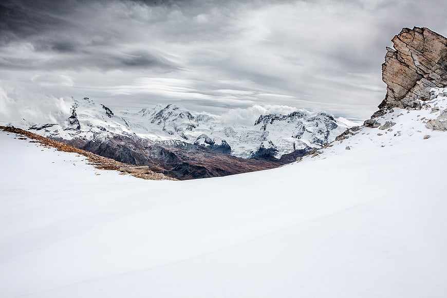 Snowy mountain landscape with dramatic clouds and rocky foreground