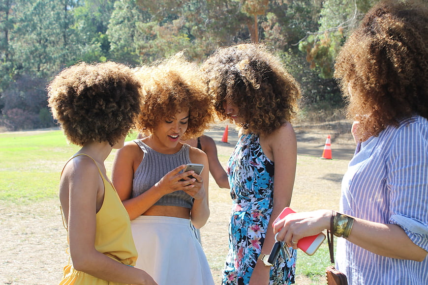 Four Women With Curly Hair Looking At Smartphone In Park