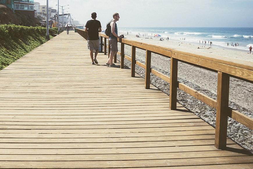 Two men walking on a sunny wooden boardwalk by the beach