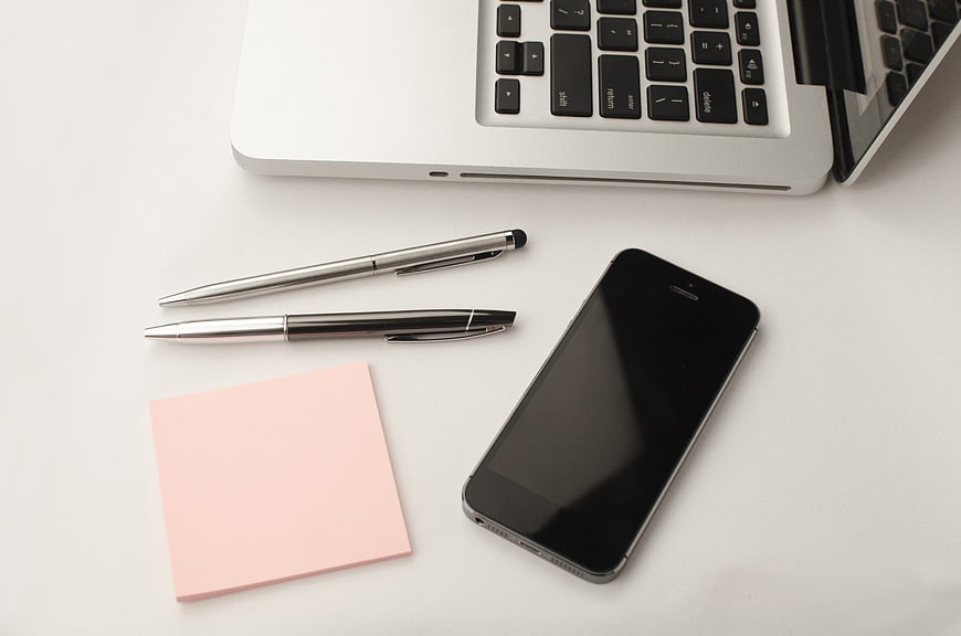 Minimalist workspace with laptop, smartphone, pens, and pink sticky note