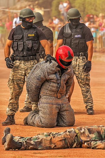 Police officers restraining kneeling prisoner in red helmet during tactical training