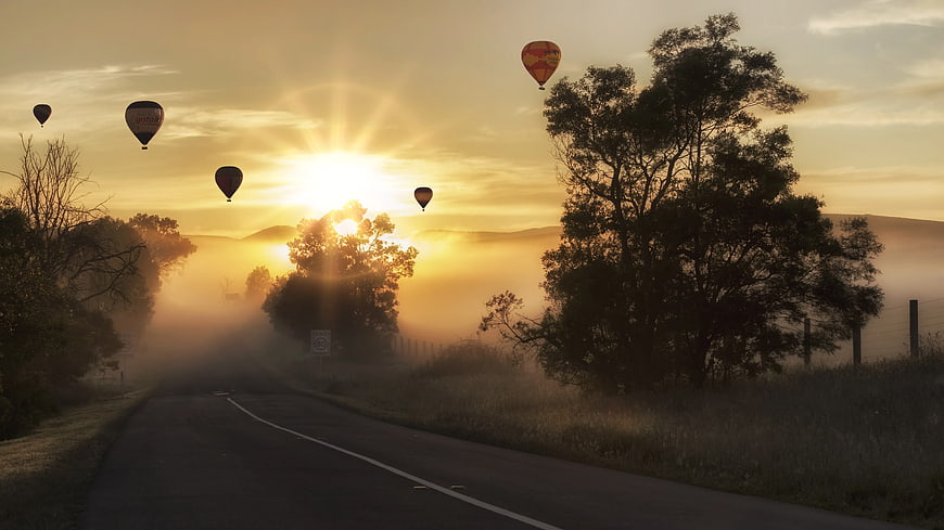 Hot Air Balloons Flying Over Misty Road at Sunrise