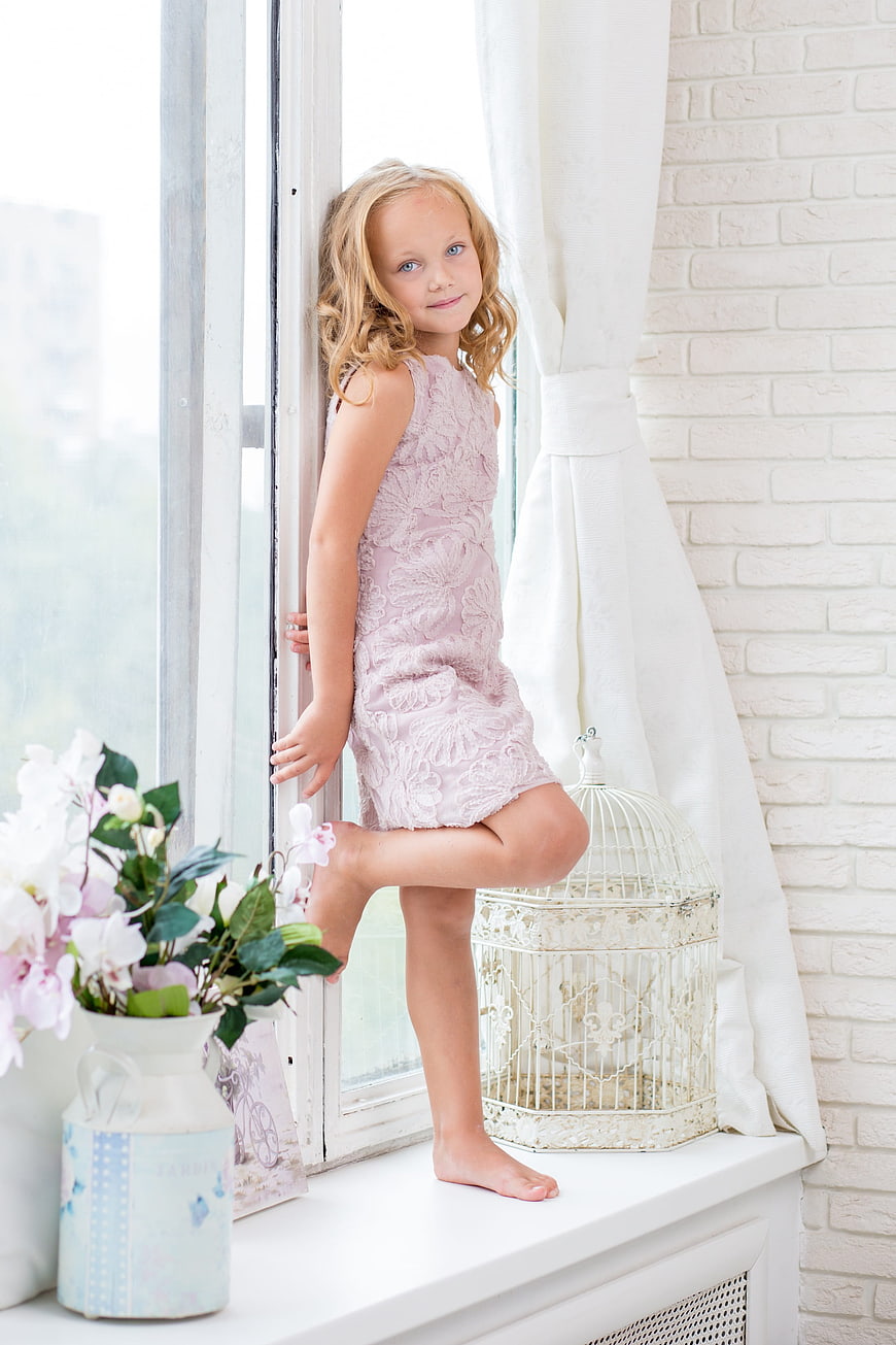 Cute young girl in pink dress standing by bright window