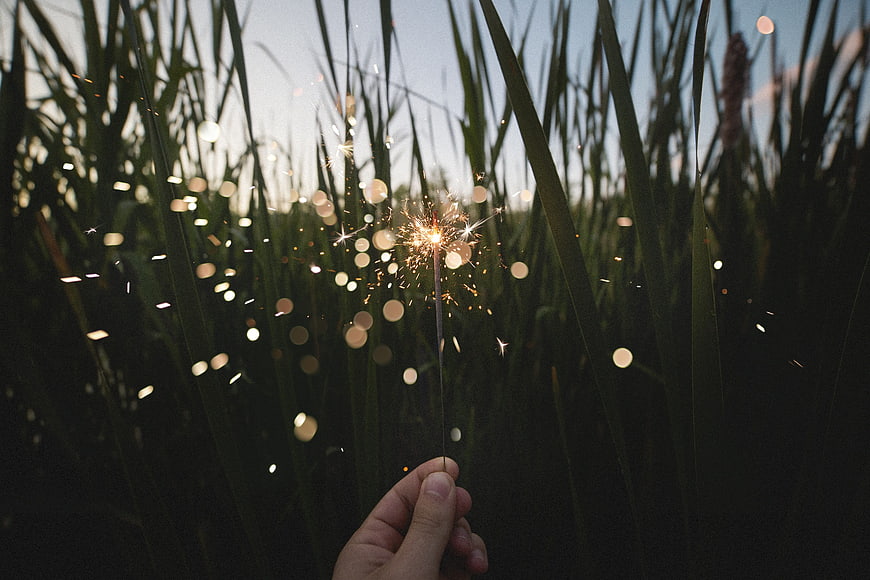 Hand holding a lit sparkler in a field of tall grass at dusk