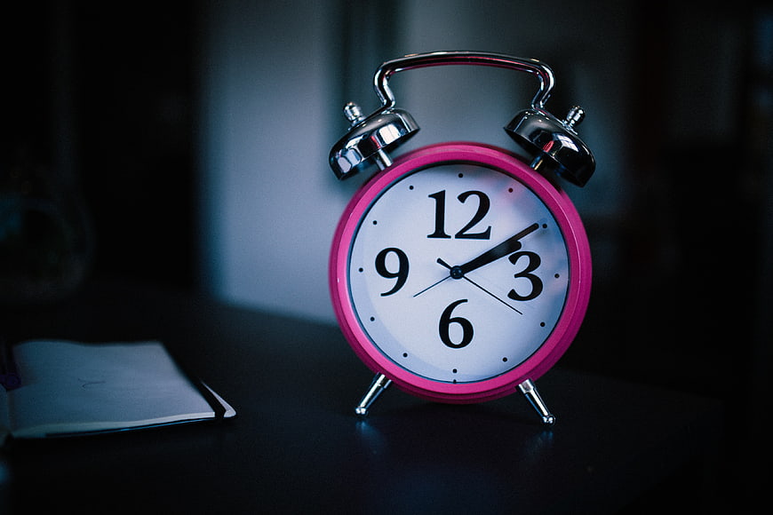Pink analog alarm clock on dark desk with notebook